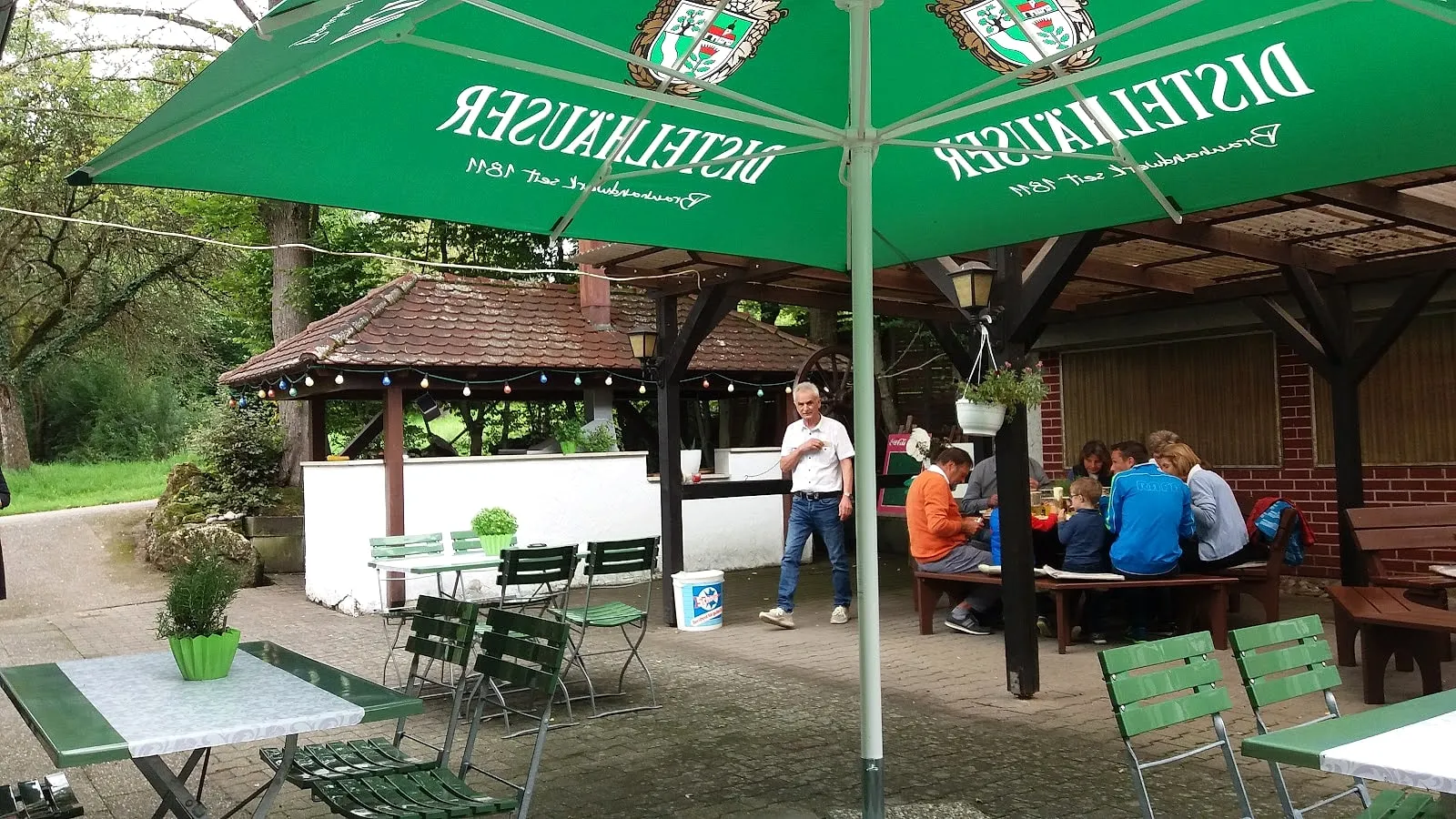 Großer Biergarten des Schützenhauses unter Bäumen, mit grünen Sonnenschirmen und Biertischen